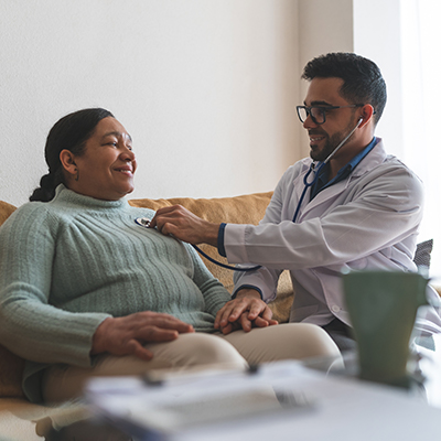 A doctor examines a middle-aged woman sitting on the sofa with a stethoscope